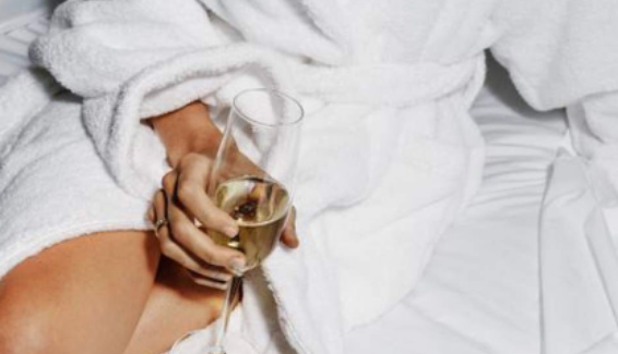 A lady in her bathrobe sitting on the bed holding a glass of champagne